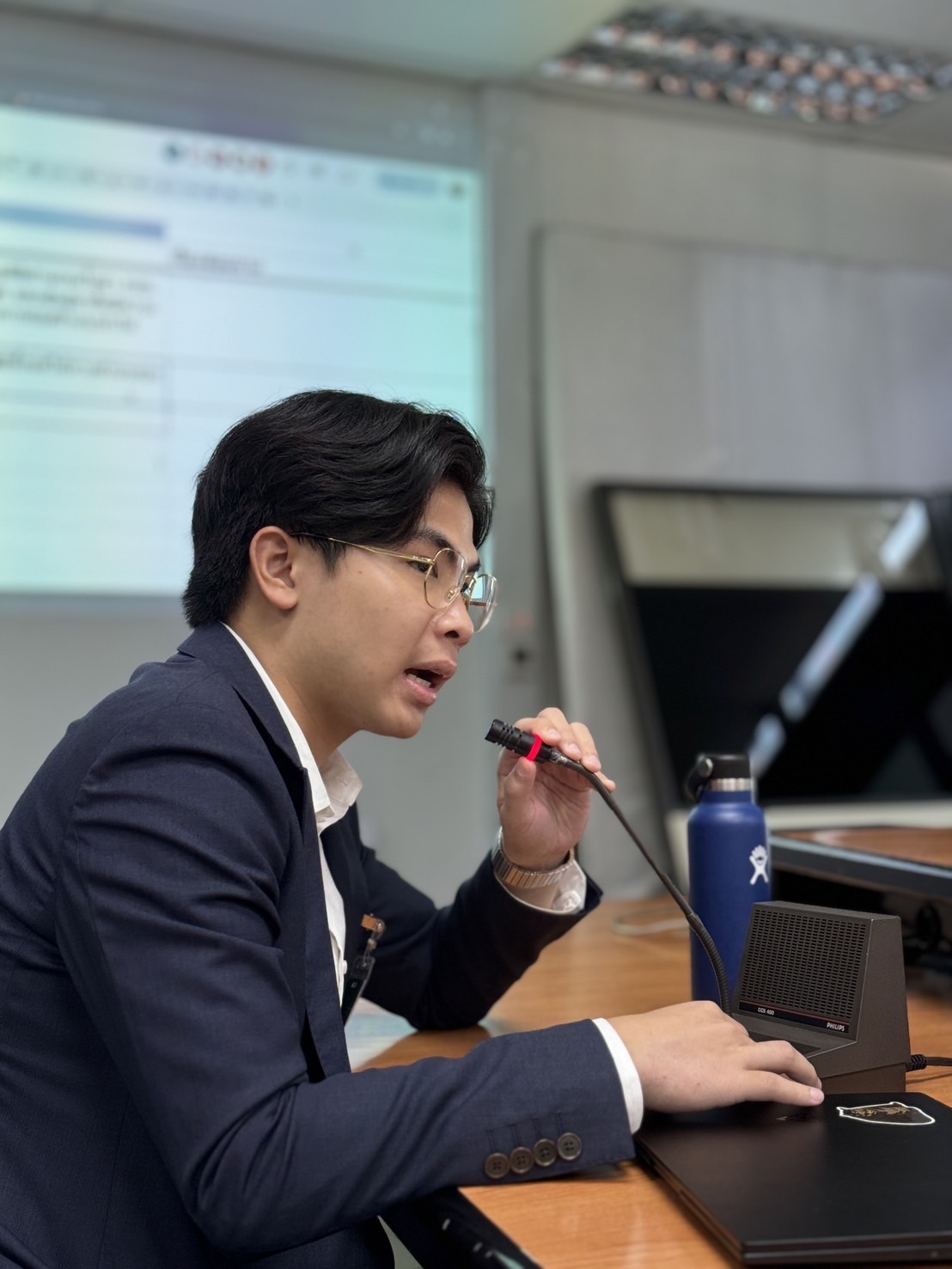 Thanapat in a navy suit speaking into a desk microphone in a meeting room